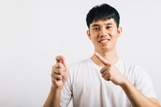 Portrait of a smiling Asian teen holding a toothbrush, brushing teeth, and pointing to it with joy. Dental health concept in a studio shot isolated on white, promoting oral care.