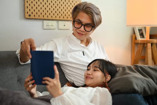 Carefree young Asian couple resting on couch at home and reading book.