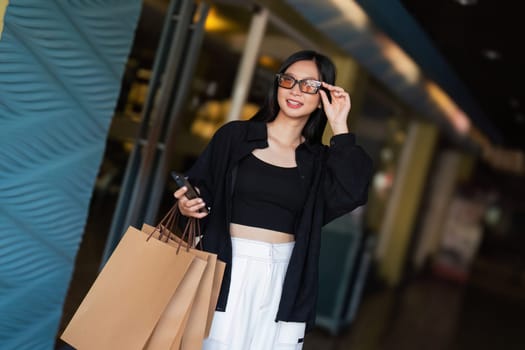 Woman using smartphone holding Black Friday shopping bag while standing on the side with the mall background.