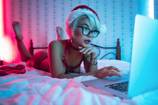Sexy girl in lingerie wearing a Santa Claus hat poses sexily on a bed with a laptop on Christmas Day in neon lights