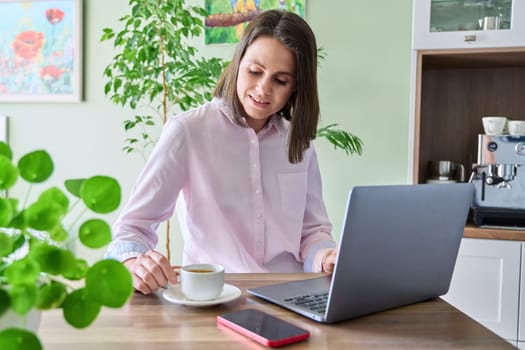 Young woman sitting at home in kitchen using laptop, drinking morning coffee. Lifestyle, freelancing, technology and work leisure, 25-30 year old people concept