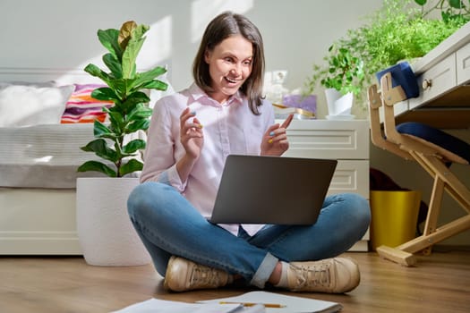 Beautiful young woman using laptop for video conference chat call. Laughing talking looking at screen female sitting on the floor at home, technology and leisure, lifestyle youth concept