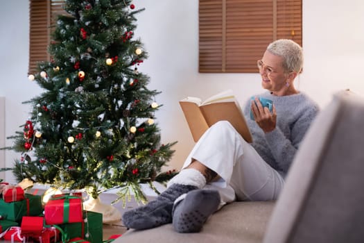 Smiling elderly woman with a cup of tea and a book relax in side of a decorated Christmas tree in living room.