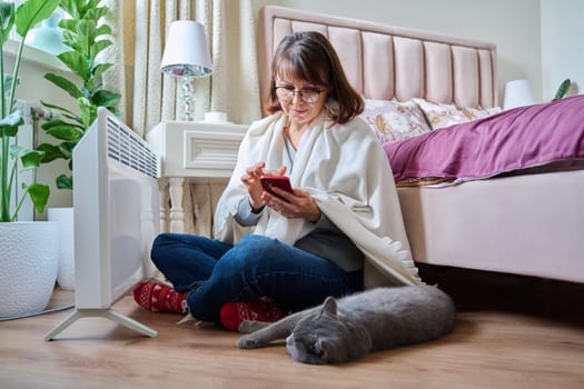 Woman with pet cat at home warming up near an electric heater. Middle aged female resting on floor, using smartphone, heating bedroom with radiator. Cold season autumn winter, comfort, energy crisis