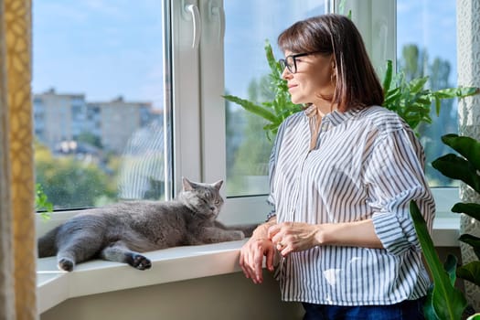 Mature smiling woman looking out window at home with relaxed cat lying on windowsill enjoying warmth of sun. Lifestyle home comfort joy happiness, animals pets, middle aged people concept