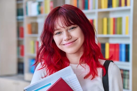 Portrait of teenage female student looking at camera in library. Smiling beautiful girl teenager with trendy red dyed hair holding notebook. College, university, education, knowledge, youth concept