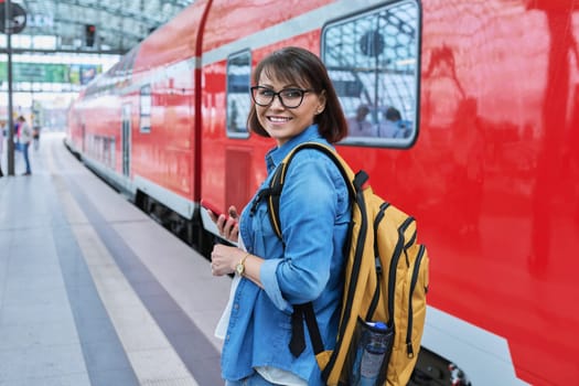 Woman waiting for railway public electric transport on platform of city station. Female with smartphone using banking app to buy online ticket payment, online timetable and route service, technology