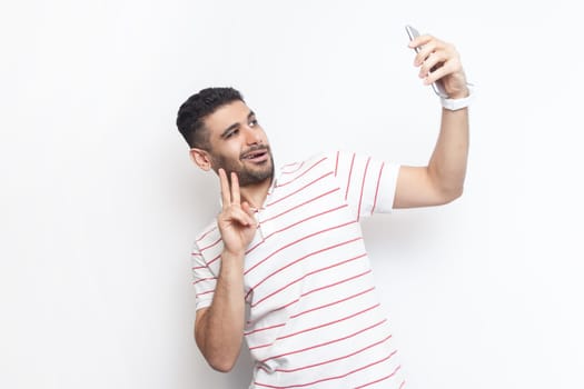 Portrait of joyful handsome playful bearded man wearing striped t-shirt standing making selfie, showing v sign to his followers. Indoor studio shot isolated on gray background.