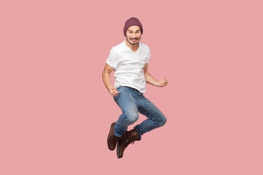 Portrait of extremely happy joyful bearded man in white T-shirt and beany hat jumping with clenched fists, rejoicing, celebrating victory. Indoor studio shot isolated on pink background.