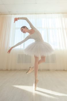 Beautiful graceful ballerina is practicing in the hall against the background of a window. Slender woman in tutu and pointe shoes in a dance class