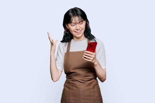 Shocked emotional young woman in apron looking surprised at smartphone screen on white studio background