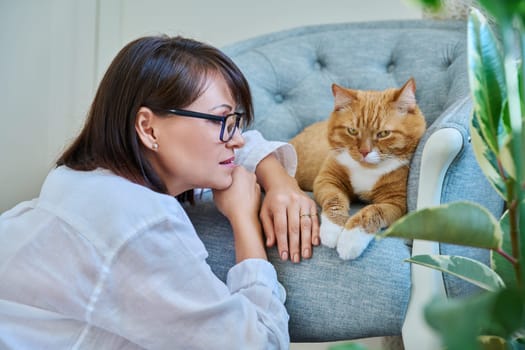 Middle aged woman talking with ginger pet cat, home interior background. Friendship, love, care, leisure, lifestyle, animals people house concept