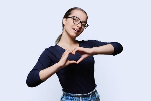 Young smiling female showing heart gesture with fingers, happy female showing love, white studio background. Body language, signs symbols, love, emotions, romance youth concept