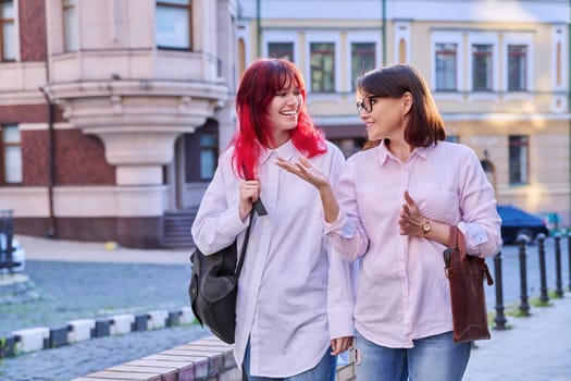 Two generations, smiling joyful happy mother and stylish attractive teenage daughter, walking talking together along city street. Family communication parent child teenager lifestyle leisure people