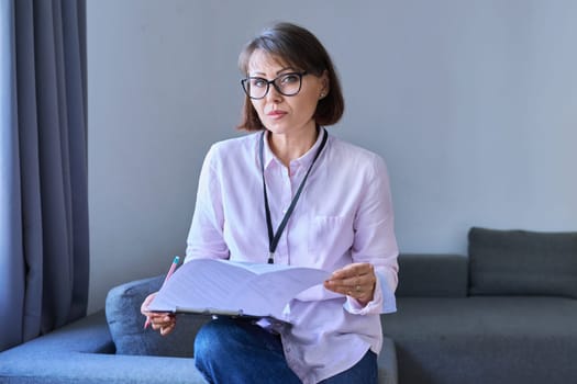 Business 40s woman with clipboard sitting on couch in office. Middle aged female advisor, accountant, agent, assistant, workplace