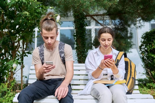 Teenage high school students, guy and girl, sitting on bench with backpacks, near academic building, using smartphones. Adolescence, youth, education, lifestyle, technology concept