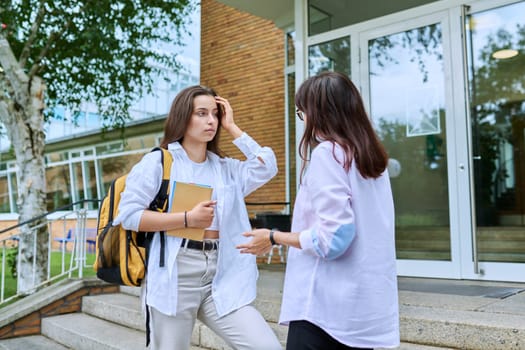 Teenage girl high school student with backpack talking to female teacher, mentor, coach, standing outdoors on steps of educational building. Adolescence, education, knowledge, communication