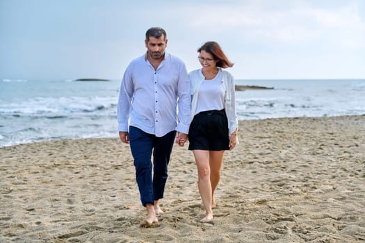 Happy middle-aged couple walking together on the beach. Mature man and woman holding hands walking along seashore. Relationship, lifestyle, vacation, tourism, sea nature, people concept