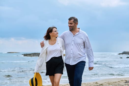 Happy mature couple walking on the beach. Smiling talking middle-aged man and woman embracing together on the seashore. Age, relationship, love, leisure, vacation, people concept