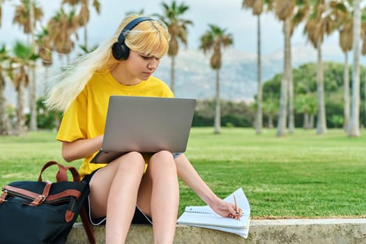 Teenage student sitting in park with laptop. Female teenager in headphones with backpack writing in notebook. Education, adolescence, high school, college, youth concept