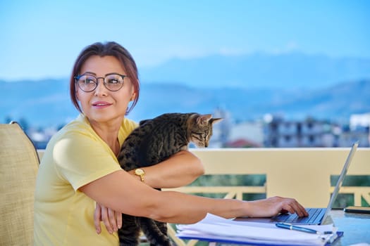 Middle-aged woman working in home office on terrace with pet cat in her arms. Remote work business, freelancing, lifestyle, animals, people concept