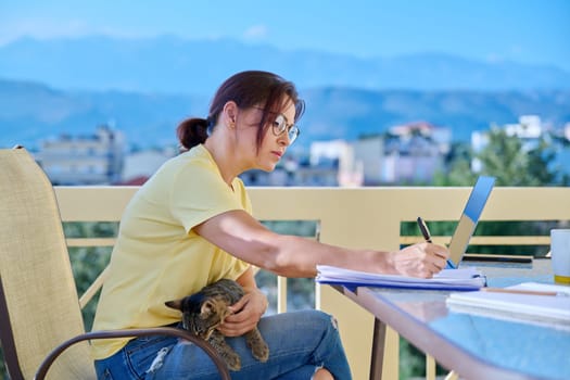 Middle-aged woman working in home office on terrace with pet cat in her arms. Remote work business, freelancing, lifestyle, animals, people concept