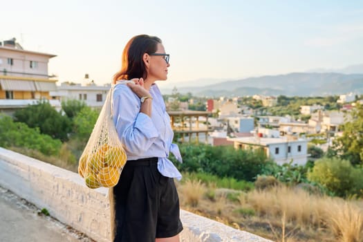 Eco tourism, woman walking relaxing in European mountain village, enjoying clean air, landscape, sunset, with a bag of fresh farm oranges. Nature, travel, ecology, lifestyle, people concept