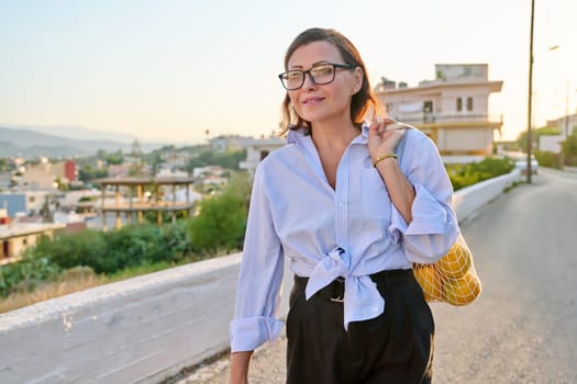 Eco tourism, woman walking relaxing in European mountain village, enjoying clean air, landscape, sunset, with a bag of fresh farm oranges. Nature, travel, ecology, lifestyle, people concept