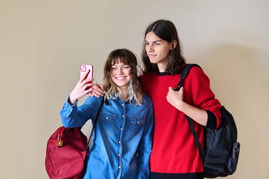 Couple of happy teenage students taking selfie using smartphone. Friends students with backpacks on the background of light wall. Youth, lifestyle, fashion, style, young people concept