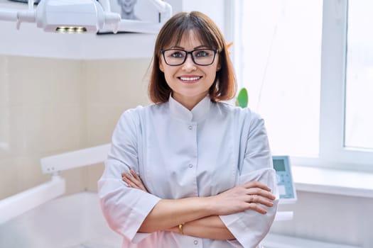 Portrait of smiling mature female dentist looking at camera in office. Confident doctor with crossed arms in clinic. Dentistry, medicine, health care, profession, stomatology concept