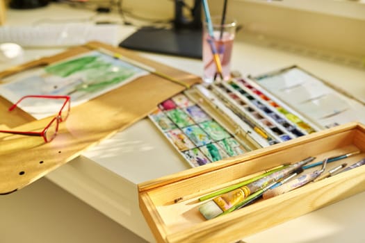 Drawing, artwork, hobby, education, creativity concept. Close-up of watercolor drawing, glasses, watercolor paints in box, brushes on the table, nobody