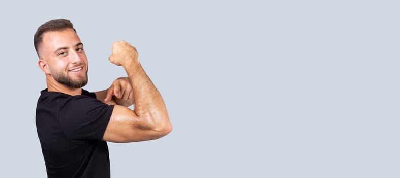 Cheerful confident strong millennial european man with beard in sportswear point finger at muscle biceps on hand, isolated on gray background. Body care, sport lifestyle and workout recommendation