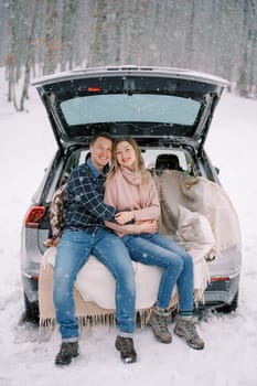 Smiling man and woman sit hugging in car trunk on blankets under snowfall in forest. High quality photo