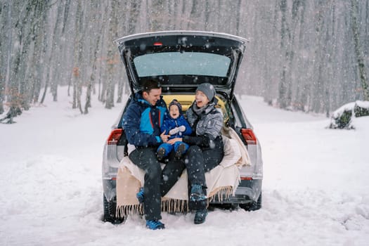 Laughing mom, dad and little girl are sitting on a blanket in the trunk of a car under a snowfall in the forest. High quality photo