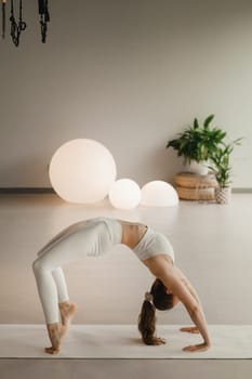 A girl in white clothes does yoga standing on the bridge on a mat indoors.