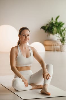 Portrait of a girl in white clothes sitting on a mat before doing Yoga indoors.