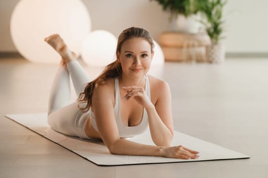 Portrait of a girl in white clothes lying on a mat before doing yoga indoors.