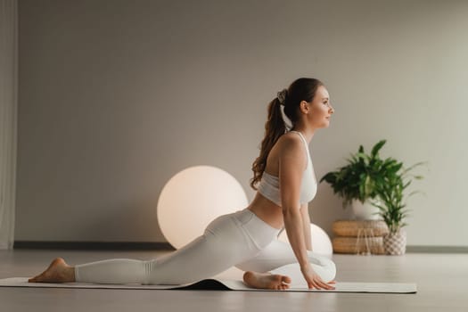 A girl in white clothes does yoga on a mat indoors.