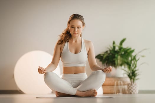 Portrait of a girl in white clothes sitting in a lotus position on a mat at an indoor yoga class.