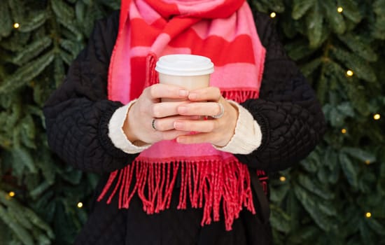 A young woman in a bright pink scarf holds a disposable glass of coffee in her hand while walking against the background of a wall of a Christmas tree.