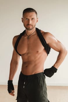 An adult muscular man with a naked torso stands in a room on a light background.