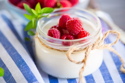Sweet cooked homemade yogurt with fresh raspberries in a glass jar, on a light wooden table.