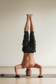 a man with a naked torso does yoga standing on his head indoors. Fitness Trainer.