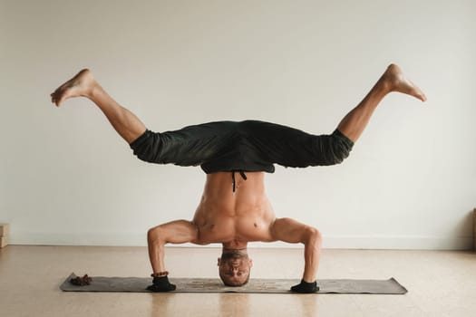 a man with a naked torso does yoga standing on his head indoors. Fitness Trainer.