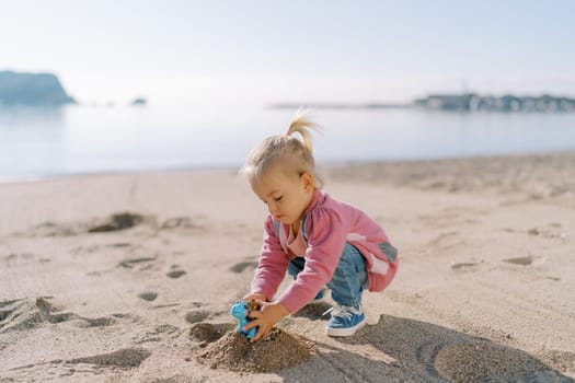 Little girl squats on the beach and pours sand into a toy dinosaur. High quality photo