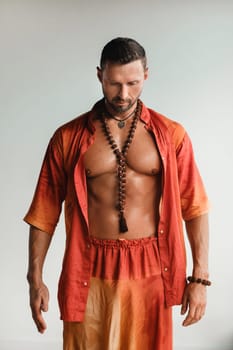 A muscular man with a naked torso in orange clothes stands in a room on a light background.