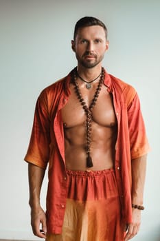 A muscular man with a naked torso in orange clothes stands in a room on a light background.