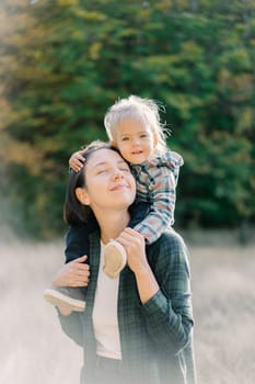 Smiling little girl hugging her mother head while sitting on her shoulders. High quality photo