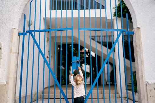 Little girl is trying to open the closed metal gate of the house while standing in the yard. Back view. High quality photo