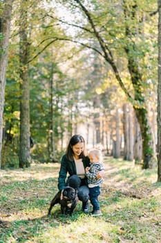 Mom pets a dog and hugs her little daughter squatting in the forest. High quality photo